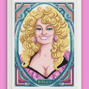 Può includere: Un ritratto a punto croce incorniciato di una persona con voluminosi capelli biondi, che indossa un top rosa con scollo nero. La figura è centrata all'interno di un'ornata cornice ovale color verde acqua. La parola DOLLY è ricamata in bianco in basso.