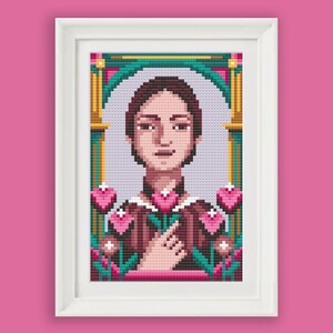 May include: Cross-stitch artwork in a white frame. The image features a pixelated portrait of a person with brown hair, surrounded by pink hearts and flowers. The background is a light purple colour, and the frame is set against a pink backdrop.