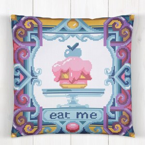 Può includere: Un cuscino decorativo con un designo ricamato colorato che presenta una torta rosa e gialla con una bacca blu in cima. La torta è su un piatto blu e bianco e le parole "eat me" sono scritte in blu su uno stendardo blu sotto la torta.