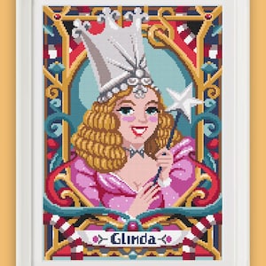 May include: A cross-stitch pattern featuring Glinda the Good Witch from the Wizard of Oz. She is wearing a silver crown and a pink dress and holding a wand. The pattern is framed in a white frame.