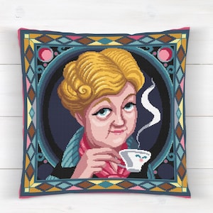 Puede incluir: Cojín cuadrado con un diseño de punto de cruz de una mujer con cabello rubio, sosteniendo una taza de té. La mujer está enmarcada por un círculo azul oscuro y un borde decorativo con patrones geométricos.