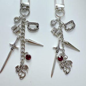 Puede incluir: Dos llaveros plateados con varios dijes. Cada llavero tiene un cierre, una gema transparente, un libro pequeño, un juego de colmillos, una espada, una gema roja y un dije de dragón. Los dijes están conectados por una cadena.