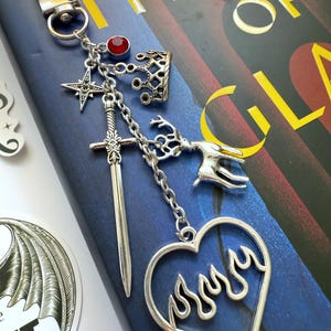 Puede incluir: Llavero plateado con varios dijes, incluyendo una corona, una estrella, una espada y un corazón con llamas. También se adjunta una gema roja. El llavero se muestra en un libro azul con el título "THRONE OF GLASS".