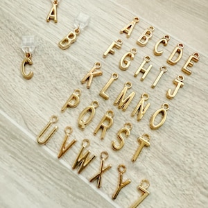 May include: Gold-colored alphabet charms, each with a small loop for attachment. The letters A, B, and C are attached to clear earring findings. The remaining letters of the alphabet are displayed on a light-colored wooden surface.