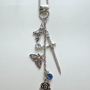 May include: Silver-coloured keychain featuring a bat, sword, butterfly, rose, and two clear and blue crystal charms. The keychain has a lobster clasp for attaching to bags or keys. The charms are suspended from a chain.