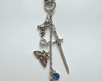 Könnte beinhalten: Silberfarbener Schlüsselanhänger mit mehreren Anhängern. Enthält eine Fledermaus, ein Schwert, einen Schmetterling, eine Rose und zwei klare und blaue Kristalle. Der Schlüsselanhänger hat einen Karabinerverschluss.