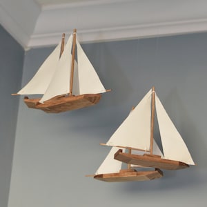 May include: Two wooden sailboat models with white sails are suspended from the ceiling. The boats are brown with white sails and are hanging from a brown circular frame. The background is a light blue wall.