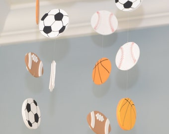 May include: A sports-themed mobile featuring various sports balls, including soccer, baseball, football, and basketball. The balls are white, brown, and orange, with black and red detailing. The mobile hangs from the ceiling against a light blue wall.