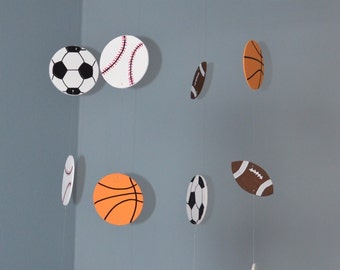 May include: A sports-themed baby mobile featuring various sports balls. The mobile includes soccer balls, baseballs, basketballs, and footballs, all suspended from a white circular frame. The balls are in their respective colors and designs.