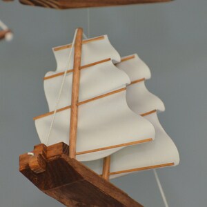 May include: A wooden model sailboat with white sails and brown wooden accents. The sails are billowing, and the model is suspended by thin white strings. The boat's hull is a dark brown color.