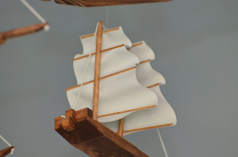 May include: A wooden model sailboat with white sails and brown wooden accents. The sails are billowing, and the model is suspended by thin white strings. The boat's hull is a dark brown color.