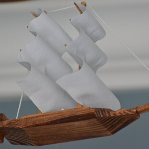 May include: A handcrafted wooden sailboat with white fabric sails. The boat is made of dark brown wood and is suspended by a thin white string. The sails are arranged in a zig-zag pattern, giving the boat a unique appearance.