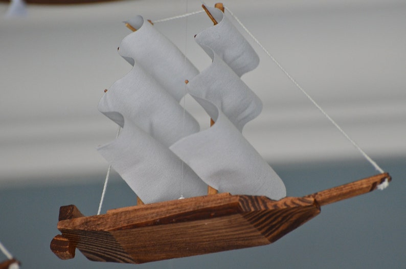 May include: A handcrafted wooden sailboat with white fabric sails. The boat is made of dark brown wood and is suspended by a thin white string. The sails are arranged in a zig-zag pattern, giving the boat a unique appearance.