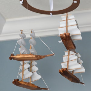 May include: A wooden ship mobile with three miniature sailboats, each with white sails and a brown wooden hull. The mobile is suspended from a brown circular frame with white ribbons attached to the ceiling.