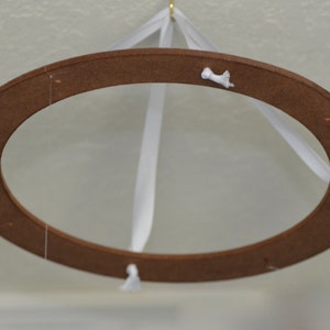 May include: A brown wooden oval-shaped mobile with white ribbons and small white decorative accents. The mobile is suspended from a gold hook, creating a delicate and airy aesthetic. The design is simple and elegant.