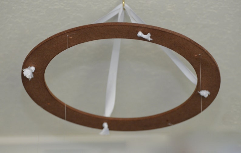 May include: A brown wooden oval-shaped mobile with white ribbons and small white decorative accents. The mobile is suspended from a gold hook, creating a delicate and airy aesthetic. The design is simple and elegant.