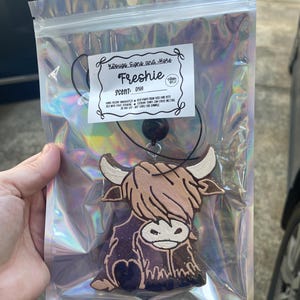 May include: A car air freshener in a clear plastic bag. The air freshener is shaped like a Highland cow with a brown and purple body, white horns, and the word "Freshie" on the label.
