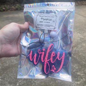 May include: A blue car air freshener in a holographic bag. The air freshener has the word "wifey" in pink glitter script. The bag also contains a label that reads "Freshie" and "Blue Hawaiian".