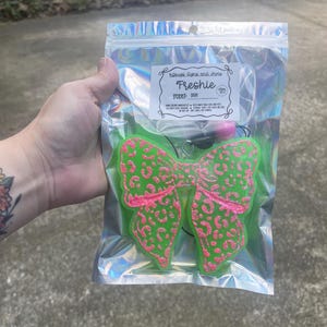 May include: A green, bow-shaped air freshener with pink leopard print accents is visible through a clear, iridescent bag. The bag has a label that reads "Freshie" and "Scent: Diva".