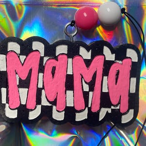 May include: A black and white checkered keychain with the word "Mama" in bright pink, outlined in black. The keychain has a silver ring and is adorned with a pink and white bead.