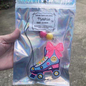May include: A blue roller skate air freshener with a pink bow and a smiley face, packaged in a holographic bag. The label reads "Freshie" and "Blue Hawaiian".