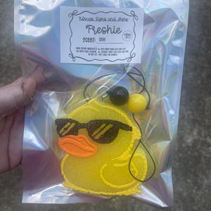 May include: A yellow duck-shaped air freshener with sunglasses and an orange beak, packaged in a clear plastic bag. The label reads "Freshie" and "Scent: Diva."