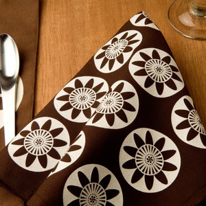 May include: A brown fabric napkin with a white floral pattern. The napkin has a repeating pattern of stylized white flowers on a brown background.