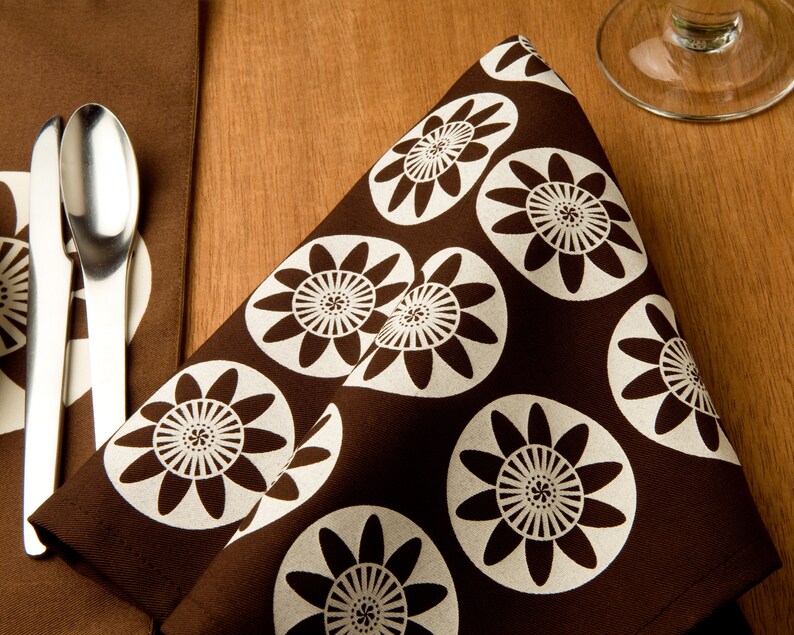 May include: A brown fabric napkin with a white floral pattern. The napkin has a repeating pattern of stylized white flowers on a brown background.