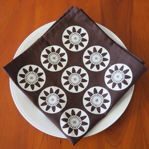 May include: A brown fabric napkin with a white floral pattern. The napkin is folded and placed on a white plate.