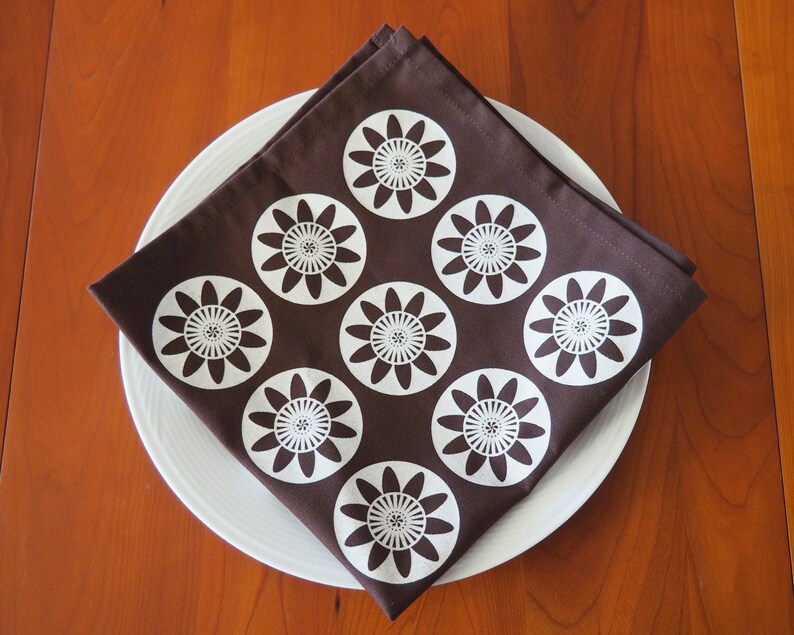 May include: A brown fabric napkin with a white floral pattern. The napkin is folded and placed on a white plate.