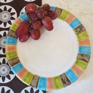 May include: A white ceramic plate with a colorful mosaic border. The border features shades of green, blue, orange, and brown. A bunch of red grapes sits on the plate.