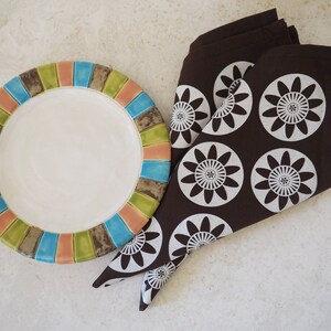 May include: A brown napkin with white floral designs is folded and placed next to a colorful ceramic plate with a white center.