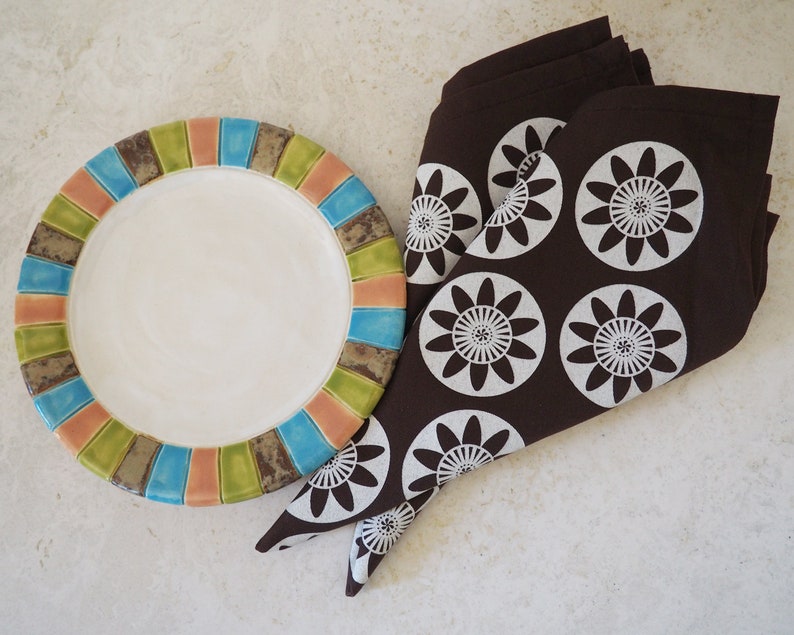May include: A brown napkin with white floral designs is folded and placed next to a colorful ceramic plate with a white center.