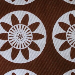 May include: Brown fabric with a repeating pattern of white stylized flowers. The flowers have a central circle with radiating lines and petals that are rounded at the ends.