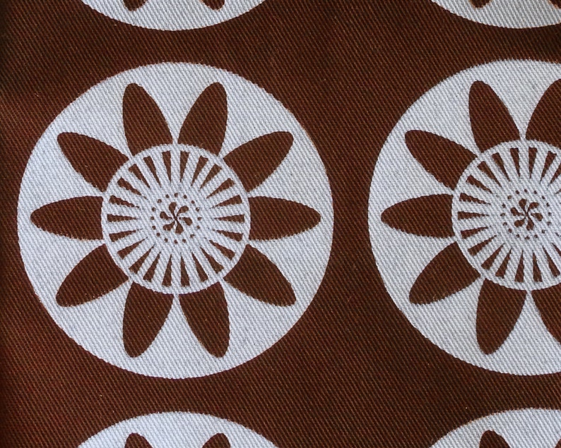 May include: Brown fabric with a repeating pattern of white stylized flowers. The flowers have a central circle with radiating lines and petals that are rounded at the ends.