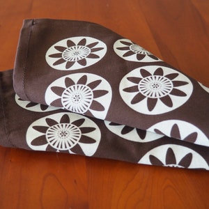 May include: Brown fabric kitchen towel with a white floral pattern. The pattern features stylized flowers with multiple petals.