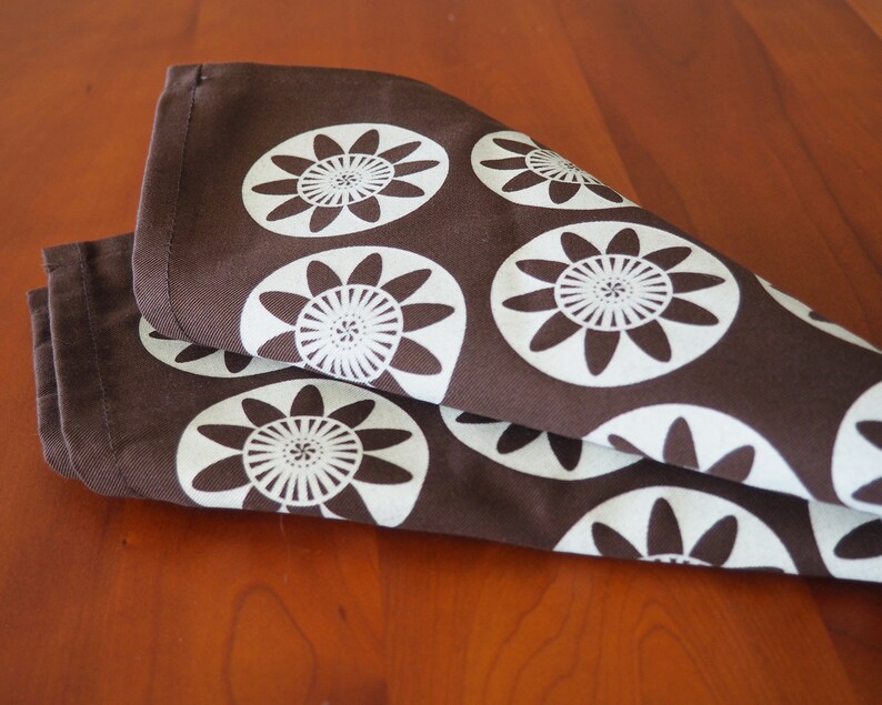 May include: Brown fabric kitchen towel with a white floral pattern. The pattern features stylized flowers with multiple petals.