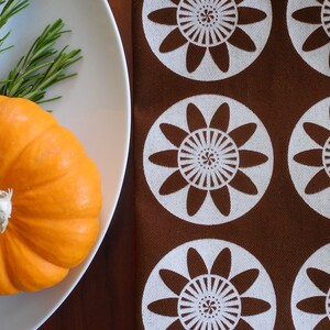 May include: A small orange pumpkin sits on a white plate with a sprig of rosemary. The plate is on a brown surface with a white and brown patterned fabric.