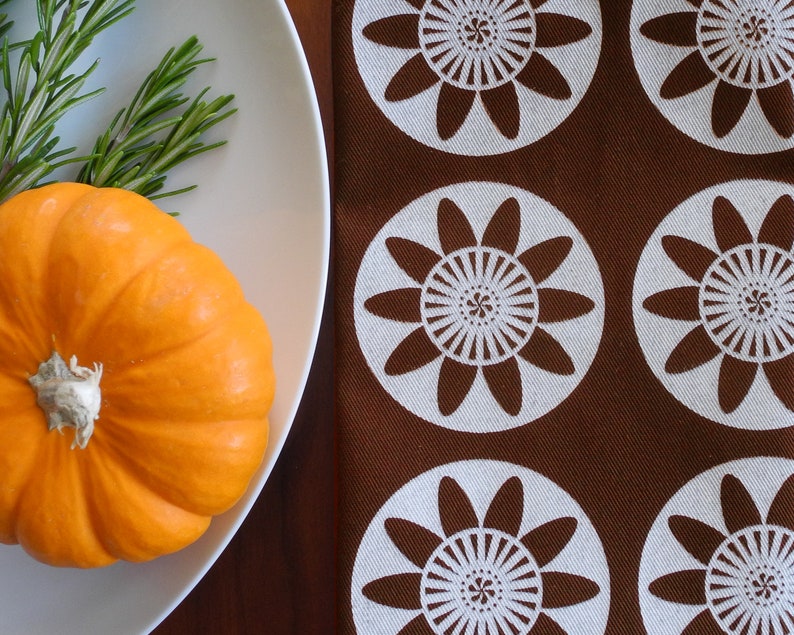 May include: A small orange pumpkin sits on a white plate with a sprig of rosemary. The plate is on a brown surface with a white and brown patterned fabric.