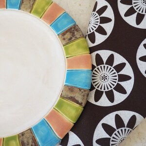 May include: A white ceramic plate with a colorful rim. The rim is made of small, rectangular tiles in shades of green, blue, pink, and brown. The plate is sitting on a brown fabric with a white floral pattern.