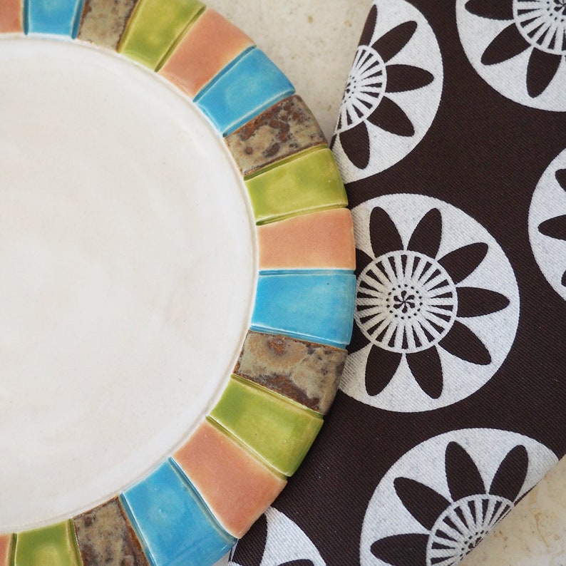 May include: A white ceramic plate with a colorful rim. The rim is made of small, rectangular tiles in shades of green, blue, pink, and brown. The plate is sitting on a brown fabric with a white floral pattern.