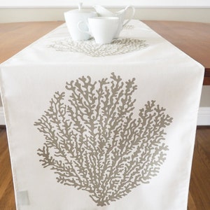 May include: A white table runner with a beige coral design. The runner is on a wooden table with white ceramic pitchers.
