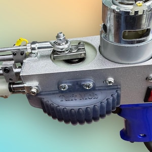 May include: Close-up of a silver and blue tufting gun. The gun features a motor, various metal components, and a blue handle. The text "Tuft Mob" is visible on a dark blue rubber grip.