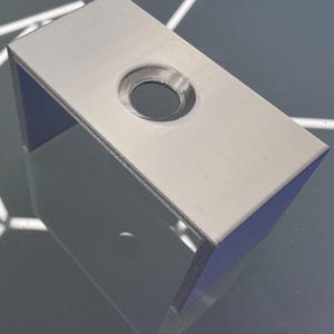 Puede incluir: Un soporte rectangular gris claro con un agujero circular. El soporte tiene un diseño en forma de U con una parte superior plana y dos lados perpendiculares. El objeto está hecho de un material sólido y tiene un acabado mate.