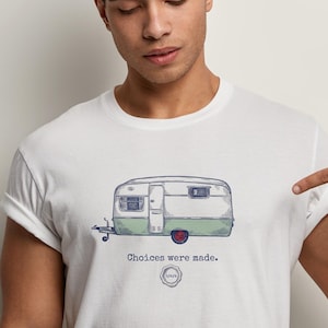May include: White t-shirt with a hand-drawn illustration of a vintage caravan in green, white, and purple. The text "Choices were made." is below the caravan. A person is pointing at the shirt.