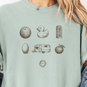 May include: A pale green t-shirt featuring brown illustrations of a coconut, duck, egg, caravan, apple, and a phone box. The shirt also has the text "Dammit Judy" and the word "Bay" in the upper right corner.