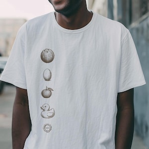 May include: A white t-shirt with a sequence of brown illustrations: a bowling ball, an egg, an apple, a rubber duck, and a target. The person in the image is wearing round sunglasses and smiling.
