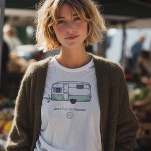 May include: A white t-shirt featuring a green and grey campervan design with the text "Zero Points Energy." The person is wearing a dark green cardigan and brown patterned trousers. The background is a blurred outdoor market.