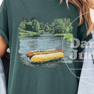 May include: Blue Spruce t-shirt with a graphic of a hot dog floating in water. The image also features a landscape with trees and a body of water. The shirt is a dark teal color.