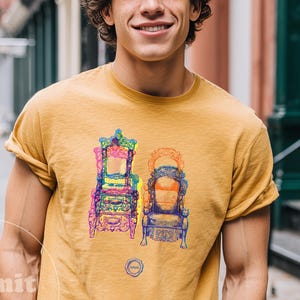 May include: Mustard-colored t-shirt featuring a colorful illustration of two ornate chairs. The graphic includes details in pink, purple, green, and blue. The shirt has rolled-up sleeves and is worn by a person in front of a city backdrop.